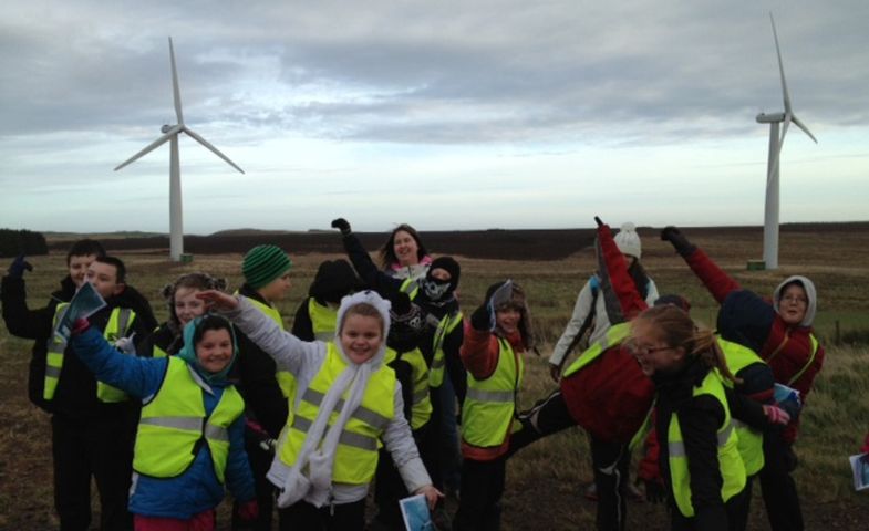 A group of children wearing yellow safety vests and speading their arms out like wind turbine blades, in front of a landscape with two wind turbines. 
