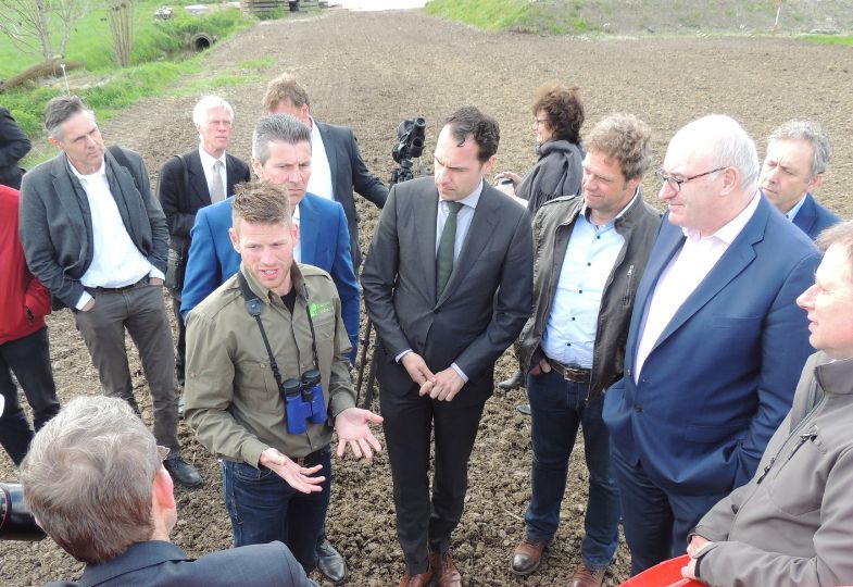 A group of people in casual clothers gathering in a field, around a politician wearing a black suit and tie.