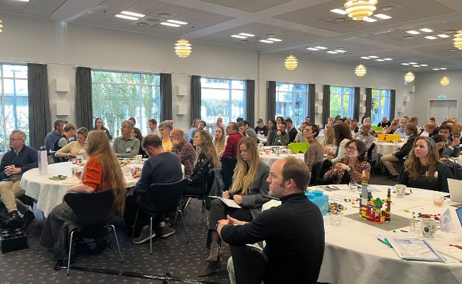 People seated at roundtables in a light room with large windows.