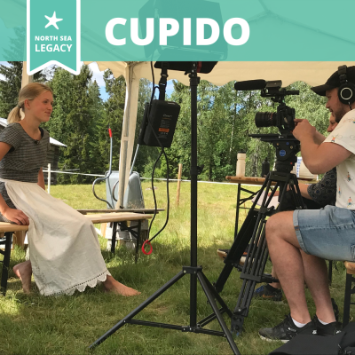 A cameraman recording a young lady wearing an apron, in an open tent on summer's day.